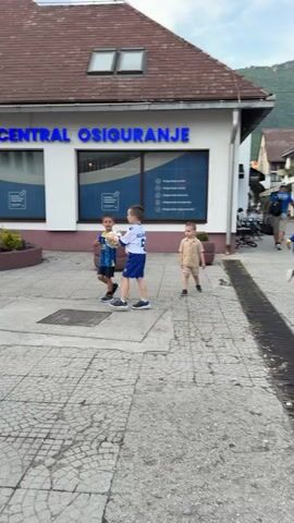 The video begins by showing a street scene in front of a building with the sign "CENTRAL OSIGURANJE". Three young boys are walking on the sidewalk. One boy is holding a soccer ball. The camera pans to the right, showing more of the street, people walking, outdoor seating for cafes, and buildings. A man wearing a black shirt and shorts walks towards the camera. Another man in a white shirt and light-colored pants walks away from the camera. Further down the street, a young girl in a blue dress is walking away from the camera. Two flags, likely national flags, are visible hanging from a building. The scene appears to be a public space in a town or city.