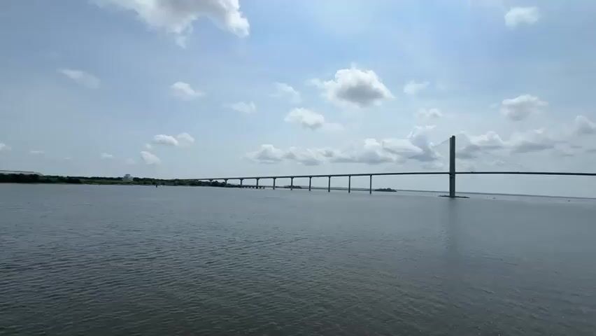 A long shot, filmed from water level, shows a wide, calm river under a cloudy sky. A very long, multi-span bridge stretches across the water, with two tall, distinct towers supporting the central span. The bridge is mostly composed of concrete pylons supporting a long roadway, with a distinctive cable-stayed section in the middle supported by the two large towers. On the right side of the frame, a tugboat is moving away from the camera and towards the bridge, creating a wake behind it. The tugboat is red and dark gray, with a black hull. The water is a muted blue-gray, and the sky is a bright blue with scattered white clouds. The far shore is visible as a thin line of green trees.