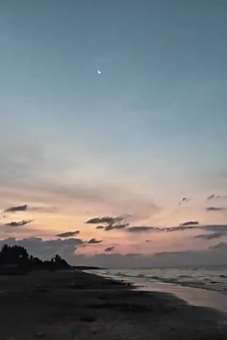 The video shows a beach at dusk or dawn. A crescent moon is visible high in the sky. The sky fades from a pale blue at the top to a mix of orange, pink, and gray near the horizon. There are scattered clouds. The beach is dark and sandy, with small waves washing ashore. Trees are silhouetted in the distance to the left.