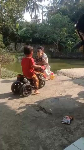 The video shows two young children outdoors. In the foreground, a child wearing a red shirt and patterned pants sits on a black three-wheeled toy motorcycle. Another child, wearing a white shirt and red pants, sits on a white toy horse with yellow wheels. The children appear to be interacting. The background shows a body of water and lush greenery, including palm trees. The scene suggests a sunny day.