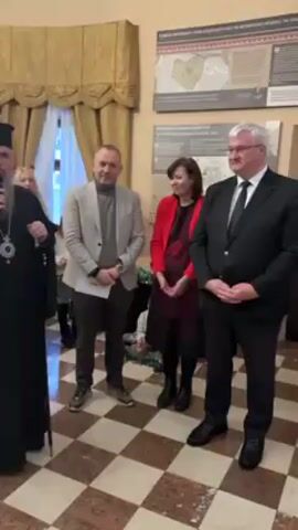 The video shows a man in religious attire speaking into a microphone. He is standing in a room with a black and white checkered floor. Next to him are two men and a woman standing. Behind the speakers are posters with information written in a Cyrillic alphabet. There is a window with gold drapes in the background.