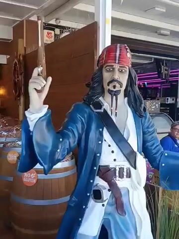A detailed, life-sized statue of Captain Jack Sparrow stands outdoors, likely at the entrance of a shop or restaurant. The statue wears a red bandana, a blue coat over a white shirt, and has long black hair, a mustache, and a braided goatee. It holds a pistol in its raised right hand. Next to the statue is a wooden barrel. In the background, the interior of the establishment is visible, featuring warm lighting, tables, chairs, and what appear to be more decorative barrels. Another person in a blue shirt and hat is partially visible in the background. The scene suggests a themed business, possibly a pirate-themed restaurant or bar, located in Puerto Del Carmen, Spain.