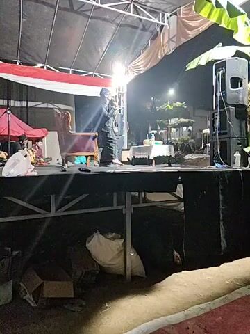 A stage is set up outdoors at night. A person is standing center stage, facing right, holding a microphone to their mouth and speaking into it. They are wearing a dark jacket over a patterned garment and dark pants. Behind the speaker, there is a large, ornate, purple armchair. To the right of the armchair, there is a small white table with items on it. Further to the right, there are two large speakers stacked on top of each other. The stage is covered by a tent-like structure with red and white fabric draped on the back wall. Beneath the stage, there are various items, including cardboard boxes and large bags. The area around the stage appears to be a public space with trees and lights visible in the background.
