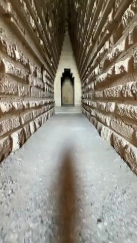 The video shows a first-person perspective moving down a narrow, stone corridor. The walls are constructed from large, stacked blocks of rough-hewn stone, with a textured surface. The corridor slopes slightly upward and tapers towards a pointed archway at the far end. The ceiling is also made of stone, with visible layers or striations. The floor appears to be a sandy or gravelly surface. A shadow of the person recording is visible on the floor, stretching ahead. The lighting is dim, casting long shadows and highlighting the textures of the stone. The overall impression is of an ancient, underground passage or tomb.