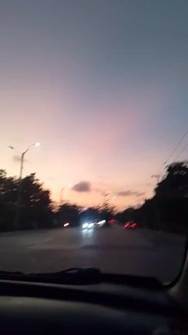 The video presents a static, forward-facing view from inside a moving vehicle, likely a car, during dusk or early evening. The sky occupies a significant portion of the frame, displaying a gradient of colors from a dark blue-grey at the top, transitioning into soft pink, orange, and purple hues closer to the horizon, indicative of a sunset or twilight. A few small, dark clouds are visible against the vibrant sky.

Below the horizon, a road stretches forward, illuminated by streetlights that are already active, casting light onto the asphalt. Power lines and utility poles are visible along the sides of the road, partially obscured by the dark silhouettes of trees. Other vehicles can be seen in the distance, their headlights and taillights glowing in the diminishing light. The scene depicts travel on a road, likely an urban or suburban street in Barranquilla, Colombia, as suggested by the location context. No people are explicitly visible, nor is any text. The weather appears clear.