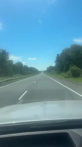 This is a first-person perspective video of driving on a highway. The car is moving at 142 kilometers per hour. The sky is clear and blue with a few scattered clouds. Trees line both sides of the road. The dashboard display shows the speed and other vehicle information, including what appears to be a route guidance system. The driver's hands are visible on the steering wheel.