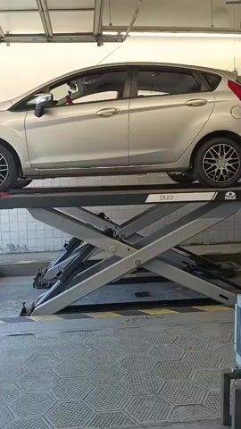 A silver car is elevated on a scissor lift inside a garage. The garage has tiled walls and a roll-up door that is partially open, revealing sunlight and the exterior. A person wearing black gloves and a yellow and blue uniform is visible on the right side of the frame, standing near the open doorway. The car lift is in the center of the frame, with the car positioned on top. The floor of the garage has a hexagonal pattern.