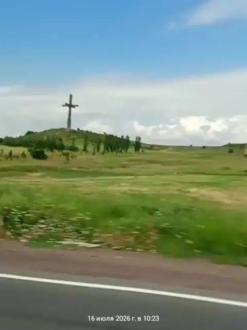 The video shows a large, dark cross on top of a grassy hill. The hill is covered in green grass and scattered trees. A line of trees is planted on the side of the hill. The sky is blue with some white clouds. The view is from a moving vehicle, as indicated by the blurred foreground and the road visible at the bottom of the frame. A timestamp at the bottom of the video reads "16 июля 2026 г. в 10:23". The overall scene is rural and peaceful.
