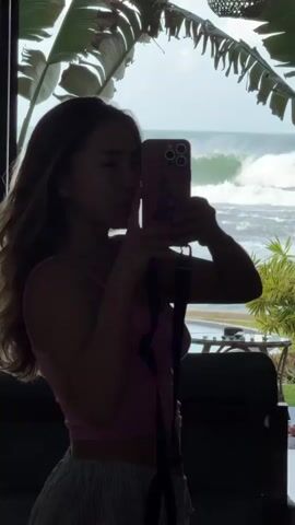 This is a vertical, medium shot video of a young woman taking a selfie in silhouette against a bright, overcast sky. She is facing right, with her head turned towards the camera she holds in her right hand. She is wearing a pink tank top and striped pants. Her left arm is bent, with her hand raised to her face, possibly adjusting her hair or posing. She has long, wavy brown hair.

The background is a dramatic ocean scene. Large, foamy waves are crashing against a dark shore. Lush green palm fronds frame the top and left side of the image, casting dappled shadows. Behind the woman, a glimpse of outdoor furniture and a railing suggests she is on a balcony or patio. The overall impression is a tropical, beachside setting. The lighting is diffused, suggesting either early morning or late afternoon, with the sun behind the camera, creating the silhouette effect. The woman appears to be in a playful mood, with a slight smile. There is no visible text in the video.