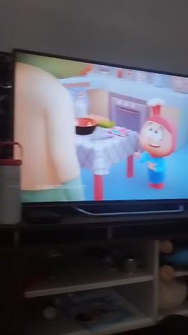 The video begins by showing a television screen displaying a cartoon. A character resembling a chef with a red hat is visible in the center of the screen. The camera then pans to the right, showing a wall, and then moves down to focus on a man sitting down, wearing a white tracksuit with black stripes. He appears to be looking to his left. The camera then pans to the right again, showing a young girl with her hair tied back in a ponytail, wearing a blue shirt. She is looking at a tablet device, which has a keyboard attached. The tablet screen shows a colorful image. Behind the girl, a guitar is visible. The environment appears to be a room with a wall and a cabinet with shelves.