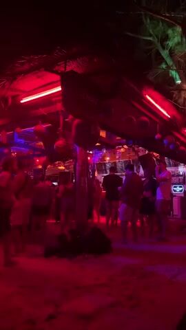 The video shows a beach party at night, heavily illuminated by red neon lights. People are standing and mingling, some are dancing to music playing. There's a bar area with a thatched roof, and the word "Chang" is visible on a sign. The ground is sandy, and some bags are lying on the ground. In the distance, lights can be seen across the water.