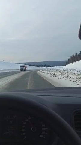 A view from inside a car shows a snow-covered landscape. The car is driving on a black asphalt road. A truck passes the car on the left. The sky is overcast. The car's dashboard is visible, displaying a temperature of 5 degrees Celsius.