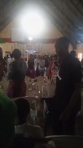 The video appears to be shot from a low angle inside a dimly lit room, possibly a church hall or community center, with a corrugated metal roof. A bright light source is visible at the top of the frame, creating a halo effect. A crowd of people, mostly obscured by shadow and motion blur, are gathered in the space. Several white plastic chairs are arranged in rows. In the foreground, a person in a dark t-shirt and light-colored pants is standing, with their back to the camera, looking towards the crowd. Other people are visible in the background, some dancing or moving. The overall atmosphere seems to be one of a gathering or celebration.