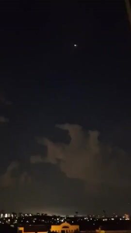 A night sky video shows a bright light in the distance, appearing to be a star or planet, above a cityscape. Clouds are visible in the sky, and the city lights below indicate it is nighttime. The light in the sky flickers slightly throughout the clip.
