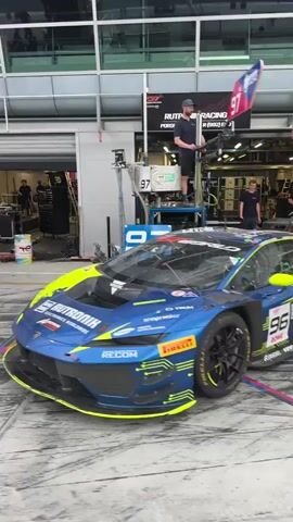 The video shows a racing pit area with multiple race cars lined up. The camera pans from a blue and yellow race car to a black and yellow race car. Some pit crew members are present. The ground is wet, suggesting a recent cleaning or rain. Above, there's a visible infrastructure of cables and overhead systems, indicating the technical and logistical setup of a professional racing environment. The pit garages are labeled with numbers. The overall atmosphere suggests preparations for or the immediate aftermath of a race.