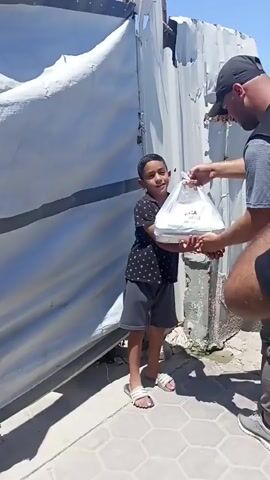 A young boy stands holding a clear plastic bag containing food items, looking at the camera. He is wearing a black t-shirt with a white pattern and grey shorts, and white sandals. To his right, a man wearing a black vest with a logo, a black cap, and grey pants is handing the bag to the boy. The background shows a damaged corrugated metal structure and a bright sky. The ground is paved with hexagonal tiles. The video appears to be taken outdoors in daylight.