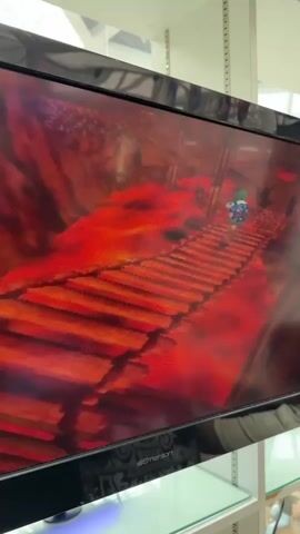 A video shows a TV screen displaying gameplay from The Legend of Zelda: Ocarina of Time. Link, the player character, is a small figure in a green tunic. He is facing a larger character with white hair, blue and purple armor, and yellow gloves. The background appears to be a reddish, cavernous environment with some red pillars. The camera briefly pans to the right, showing a different section of the game environment with more red pillars and a brown floor.
