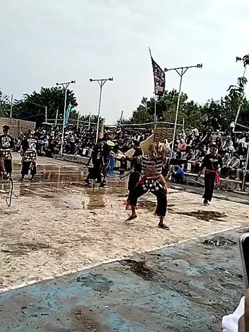 The video captures an outdoor performance with a crowd gathered to watch. In the foreground, several individuals are engaged in a dance or martial arts demonstration. One performer, positioned centrally, is wearing striped attire and a headdress, striking a dynamic pose with arms extended. Other performers in dark clothing are visible on the left side of the frame, some holding what appear to be staffs or poles. The performance area is a flat, wet surface, possibly made of concrete or dirt, with puddles of water reflecting the overcast sky. In the background, a tiered seating area is filled with spectators of various ages, most of whom are looking towards the performance. Two tall poles with lights or speakers are erected behind the performance area. A flag with an emblem is visible flying from one of the poles. The overall environment suggests a public gathering or festival.
