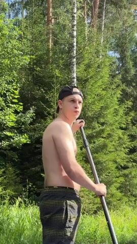 This video shows a young man stand-up paddleboarding on a calm river in Siversky, Russia. The man, wearing a black baseball cap and checkered shorts, is shirtless and holding a paddle. He is positioned on the paddleboard, facing towards the right side of the frame, with his paddle in the water. The paddleboard is blue and white with a colorful abstract design.

The scene is set in a lush, green environment with tall trees, likely coniferous, forming a dense backdrop. The river is clear and reflects the surrounding greenery. The water is calm, with some aquatic plants visible beneath the surface. The lighting suggests it is daytime, likely late morning or early afternoon, with bright sunlight filtering through the trees. The overall mood is peaceful and relaxed, conveying a sense of enjoyment of nature. There is some text visible on the paddleboard, including "R-Lite".