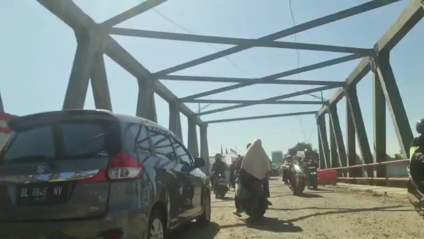 The video shows a gray Suzuki Ertiga car driving across a metal bridge with a rough, gravel-covered surface. The car is moving slowly, and several motorcycles are also traversing the bridge ahead. Many of the motorcyclists appear to be women wearing hijab. The bridge structure is composed of interlocking metal beams. The weather is sunny, and the sky is clear. A barrier of red and white stripes is visible on the left side of the bridge. The car's license plate reads "BL 1846 NV."