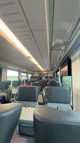 The video shows the interior of a passenger train car. The seats are upholstered in black leather and are arranged in a forward-facing configuration. The aisle is visible between the rows of seats, and overhead luggage racks are present. Natural light enters through the windows, and artificial lighting is provided by long, illuminated panels on the ceiling. The environment appears clean and well-maintained. There are a few passengers visible, seated and looking forward. The train is in motion, as indicated by the slight blurring of the exterior scenery visible through the windows.