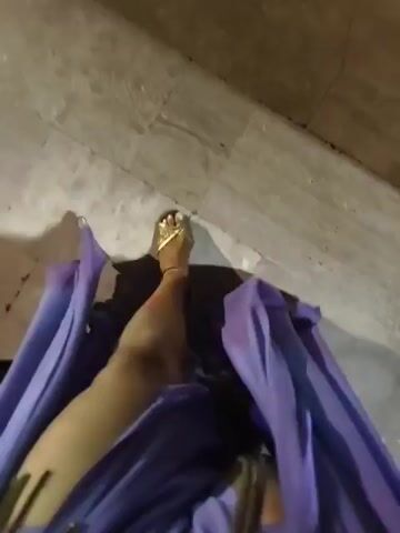 This video captures a first-person perspective of someone walking down a tiled staircase at night. The staircase is flanked by a white railing on the left. At the bottom of the stairs, a brightly lit swimming pool area is visible, with lounge chairs, tables, and a building in the background. The building appears to be a hotel, with multiple floors and lit windows. There are people seated at tables near the pool, suggesting an outdoor dining or lounge area. Pink flowers are blooming on the right side of the frame, adding a touch of color to the scene. The overall impression is a relaxed evening atmosphere at a resort. The lighting suggests it is nighttime. There is no visible text.
