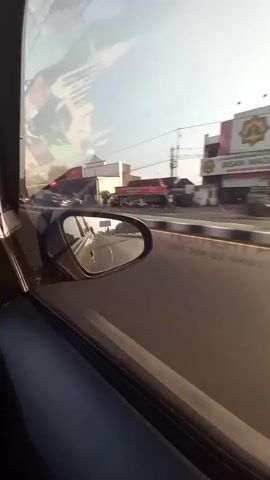 The video shows a view from inside a car, looking out the side window and the side mirror. The car is moving along a road lined with buildings and trees. In the side mirror, other vehicles can be seen. The sun is shining, and the sky is clear.