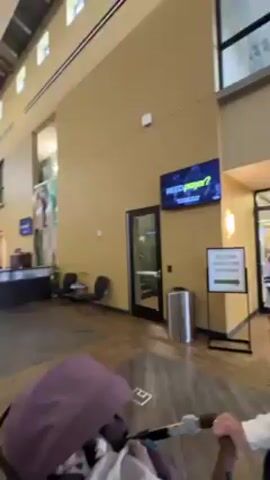 The video shows an interior shot of a large foyer, possibly the entrance to a fitness center or community center. The camera is being held by someone who is walking and filming. Initially, a man in a white shirt is visible, greeting someone off-screen with “Good morning.” The camera then pans to show the vastness of the space. There are several people scattered throughout the area, and a receptionist desk is visible on the right. A woman wearing a mask is standing in the middle of the room, cheerfully welcoming people with open arms. The space features high ceilings, large windows, and neutral-colored walls. The floor is a brown wood laminate. There are black cocktail tables in the background.