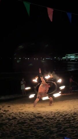 The video shows a shirtless man with tribal tattoos wearing tribal shorts performing a fire dance on a sandy beach at night. He is using a long, thin pole with flames on both ends, twirling it expertly around his body. A few people are standing in the background, one of them holding a phone. Small bonfires are visible on the beach. Colorful flags hang overhead on a string. In the distance, lights from a building can be seen across the water.