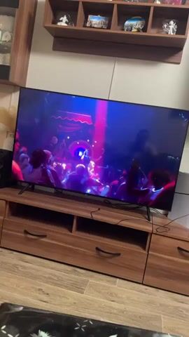 The video shows a television screen displaying various scenes. The first scene shows a crowded nightclub with people dancing under blue and purple lights. The second scene is an aerial view of a bridge over a bright blue ocean, with a small airplane in the sky. The third scene depicts a woman in a white bikini posing by a swimming pool with a city skyline in the background. Another woman in a red swimsuit is visible in the pool. The video appears to be a montage or trailer.
