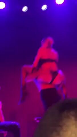 Here is a description of the video:

The video shows a stage performance with two shirtless men and a woman in lingerie. The woman is wearing a black bra and thong, with thigh-high stockings. The men are in black pants, one wearing an armband. They are dancing together under purple and white stage lights. One of the men sits on a chair while the woman dances around him. The view is from the audience, with the back of someone's head visible in the foreground.