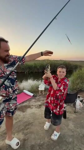 The video shows a young boy proudly holding up a caught fish. He is wearing a red shirt with a floral pattern and dark shorts. He is standing next to a body of water, likely a river or a pond, surrounded by tall grass. A fishing rod is visible, and another person, partially visible on the left, is also present. The sun is setting in the background, casting a warm glow over the scene. There are some camping items, including a pink mat and a chair, scattered around. The boy seems to be enjoying his fishing experience.