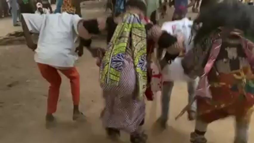 The video shows a group of people dancing in a dusty outdoor area. Several of the dancers are wearing costumes, including one with a large, shaggy, dark wig and another with a colorful patterned garment stuffed to create the impression of a large belly. Other dancers are wearing casual clothing such as white shirts and colored pants. The dancers are surrounded by onlookers. The atmosphere appears festive, with sounds of music and celebration in the background.