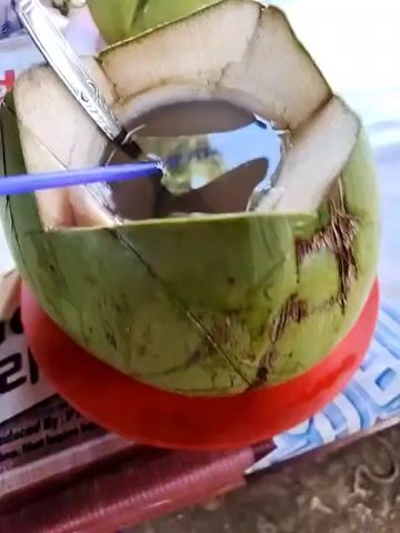 The video shows two young coconuts on a table. One is open, with a spoon and straw sticking out, and sits on a small red plate. The other is whole, sitting on another small red plate, with a straw sticking out. A person's arm is visible in the background, and a motorcycle is visible through the window. The table has a printed advertisement on it.