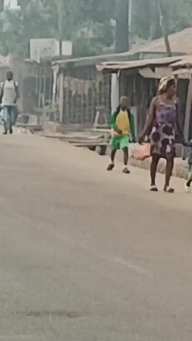 The video shows a street scene in a rural or developing area. The road is paved but looks worn. There are simple, somewhat dilapidated buildings lining the street, constructed with wood and corrugated metal. A woman in a purple patterned dress walks with two young children, one in a blue patterned shirt and one in an orange and white shirt. A slightly older child in a yellow shirt and green shorts walks ahead of them. In the background, a man in a white shirt walks in the same direction as the woman and children. A man on a motorcycle passes in the opposite direction. The light suggests it is early morning or late afternoon, with a hazy or muted atmosphere. The overall impression is of a community with limited resources in a relatively undeveloped setting.