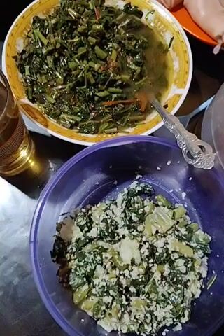 The video shows two dishes with food. One is a purple bowl filled with chopped green vegetables and white cheese, seemingly mixed. The other is a yellow and white plate filled with cooked leafy green vegetables with a thin broth, being stirred with a spoon. The scene appears to be a dinner table.