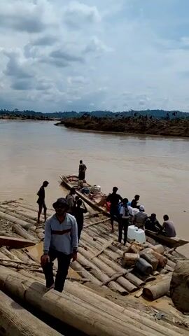 The video depicts a muddy river landscape. Several people are visible on what appears to be a makeshift wooden raft or platform constructed from logs. A smaller boat is also present, loaded with supplies, and people are either on it or getting on it. The raft or platform leads to a muddy riverbank where a group of people are standing. The river itself is wide, murky, and seemingly overflowing, with debris visible in the water and along the banks. The sky is overcast with clouds.