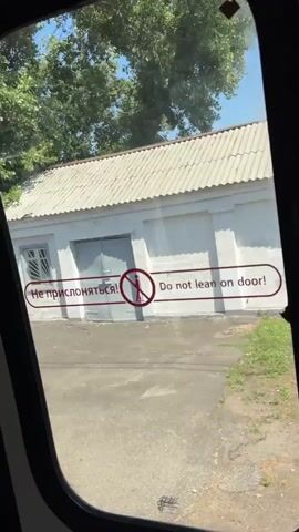 A video captured from inside a vehicle shows a view through a window of a building and surrounding environment. The building is white with a grey roof and appears to be a garage or storage structure with two large doors. A red and white sign with a prohibition symbol and text in two languages, "Не прислоняться!" and "Do not lean on door!", is visible on the doors. Trees and foliage are present behind the building. The ground in front of the building is paved. The video appears to be taken while the vehicle is moving, as evidenced by the slight blur and changing perspective of the scenery. The overall impression is a mundane, everyday scene observed from a moving vehicle.
