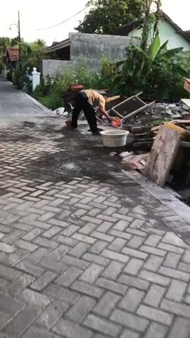 The media file captures an outdoor scene in Gelam, Indonesia, likely during daylight hours given the bright illumination and shadows. A person, dressed in dark pants and a patterned shirt, is actively engaged in a task. They are bent over, interacting with a large sieve-like object supported by a wooden frame. To the person's right, a large, light-colored circular tub sits on the ground, and a smaller orange bucket is nearby.

The ground is paved with interlocking bricks, exhibiting a wet or dark sheen in places, suggesting recent activity or possibly residual moisture. In the background, elements of a residential setting are visible. There is a building with a light-colored facade and what appears to be a green-roofed structure behind it. Lush green vegetation, including banana plants, flourishes in the area, contributing to a tropical feel. Piles of materials, possibly construction-related, are also present, including stacks of reddish tiles and debris. A white decorative post is also visible on the left side of the frame.

The overall impression is one of work or preparation, possibly related to construction or a crafting process. The individual's posture and focus suggest concentration on the task at hand. The environment appears to be a semi-urban or rural area, typical of Indonesian villages. There is no visible text in the media file.