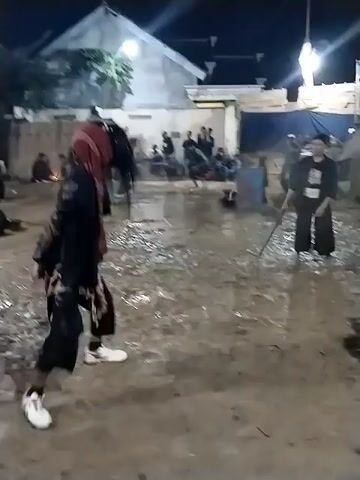 At night, an outdoor scene in Jenggrik, Indonesia, shows several men engaged in a dynamic cultural performance or martial arts display on a muddy ground. The area is lit by artificial lights, including streetlights and lights on buildings.

In the foreground, a man is seen from behind, wearing a dark long-sleeved shirt with a logo and a patterned cloth around his waist. In the mid-ground, another man in a black outfit, a red head covering, and white sneakers performs a crouching stance. Other participants are actively moving across the muddy terrain, some falling and getting up. One man is briefly seen lying in the mud before getting up.

In the background, temporary structures, possibly fences or stalls, are visible, along with a white building. A group of spectators is gathered behind these structures, observing the event. White flags are hanging from poles. The ground is thoroughly soaked and muddy, suggesting recent rain or intentional watering for the performance. The overall atmosphere appears to be a communal gathering for this traditional display.