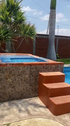 The video shows a swimming pool area. The pool is rectangular and filled with clear blue water. There are steps leading into the pool, and a small jacuzzi or hot tub is attached to the pool's side. The surrounding area is paved with light-colored tiles. Palm trees and other plants provide landscaping around the pool. A brick wall with a fence runs along the back of the property. The sky is partly cloudy, indicating a sunny day. The camera pans slightly to show more of the pool and surrounding landscape. The scene is peaceful and suggests a relaxing atmosphere.