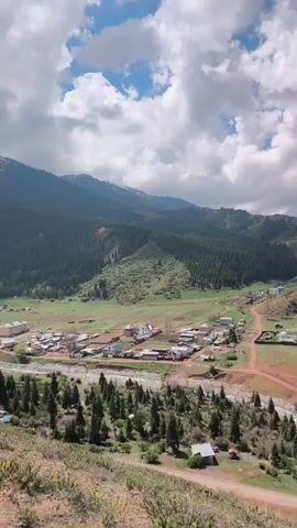 The video presents a scenic, high-angle panoramic view of a valley in Jeti-Ögüz, Kyrgyzstan, under a bright sky with scattered white clouds. The camera slowly pans from left to right, revealing the expansive landscape.

Initially, the left side of the frame shows a small village or settlement comprising several houses with varied roofs, nestled amidst green fields and sparse trees. A wide, stony riverbed, possibly dry or with low water, flows through the valley, separating parts of the village. Beyond the settlement, lush green, forested mountains rise, forming the backdrop.

As the camera continues to pan, it unveils the dramatic red rock formations characteristic of the Jeti-Ögüz region. These striking, layered red cliffs dominate the right side of the valley, featuring rugged textures and varying shades of red and orange. At the base of these formations, more green areas, pine trees, and a few larger buildings (possibly part of a resort or complex) are visible near the river. A dirt road winds through the valley floor, and a white car or van can be seen driving along it, providing a sense of scale. The overall impression is one of tranquil natural beauty, highlighted by the vibrant contrast between the red rocks and the surrounding greenery. No visible text is present in the video.