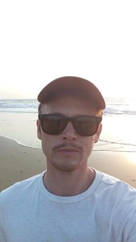 A man wearing a brown cap and sunglasses stands on a sandy beach with the ocean behind him. He is wearing a light-colored t-shirt and has a mustache. The sun is shining, casting a bright light on the scene. The sky is blue, and waves are visible in the ocean. The camera appears to be held by the man, capturing a selfie. The angle of the camera changes slightly to show more of the sky.