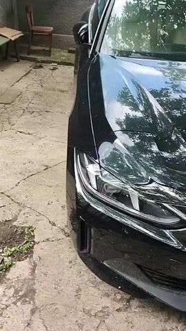 The video pans across the front of a black BMW car. The car is parked on a cracked concrete surface with some green foliage visible around the edges. The car's headlights are on, and its grille is prominently displayed. A circular decal with the letters "PM" in a stylized font is affixed to the hood of the car. The license plate is visible and reads "AI 3792 YB". A wooden chair and a pile of debris are visible in the background to the left. The overall impression is a static shot of the car, possibly being presented or showcased.