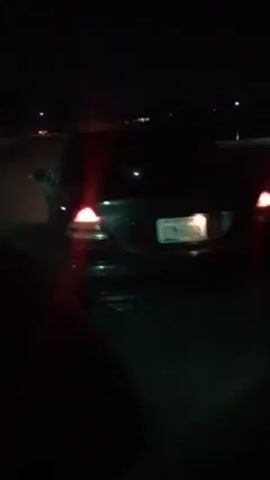 The video shows the rear of a black sedan at night. Its taillights are illuminated, and the license plate is partially visible. The camera then pans to the side, showing a green beam of light moving across the road. The road surface is visible, and there are some distant lights in the background. The video is dark and shaky.
