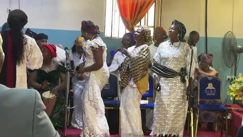 A group of women in traditional clothing are standing in a room. The woman on the far right is wearing a white dress and a black and white striped sash. The woman to her left is wearing a white dress and a brown and gold patterned shawl. The woman in the center is wearing a white lace dress with ruffled sleeves and a red beaded necklace. The woman to her left is wearing a white dress and a red headscarf. Behind them, another woman is playing a drum. The room appears to be a church or a similar gathering space, with chairs and a microphone visible. The overall atmosphere seems festive.