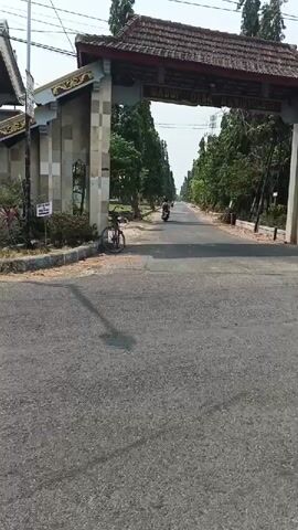 A video shows a street scene with an archway in the background. An overcast sky is visible. The archway has the words MASUK DESA SUMBER KECIL written on it. Two motorbikes are visible on the road, one in the distance and another closer to the archway. A bicycle is parked on the left side of the road near the archway. In the foreground, the back of a three-wheeled vehicle with a covered cargo area is seen moving away from the camera. Trees line both sides of the road.