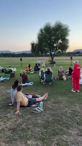 A video captures an outdoor gathering with a group of people seated on the grass, attending a performance. In the center, three individuals are performing under a large tree: one playing a keyboard, one singing, and one playing a guitar. Two speakers are positioned on either side of the performers. Spectators are seated in various chairs and on blankets, facing the performance area. The environment is a grassy field with trees and hills in the background, suggesting an evening or late afternoon setting with the sky transitioning to dusk. The overall scene depicts a relaxed, informal concert or event in a natural setting.