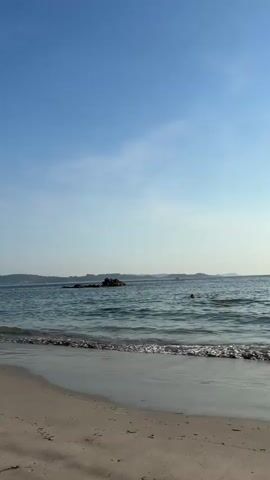 The video shows a tranquil beach scene with a clear blue sky. Gentle waves lap against a sandy shore in the foreground. In the distance, there is a small rocky island with some vegetation. A few boats are visible on the horizon. The water is a shimmering blue-green color. The overall atmosphere is peaceful and serene.