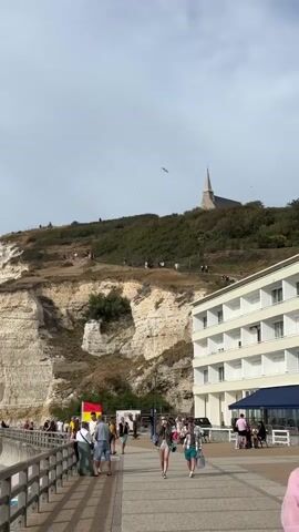 A wide-angle vertical video shows a crowded promenade beside a white building. People are walking in both directions on the promenade, which has a railing on the left. In the background, a steep, grassy cliff rises with a winding path leading to a church at the summit. Several people are visible walking up the path. The sky is overcast with some birds flying. A red and yellow flag is visible on the promenade. The building appears to be a hotel with balconies.
